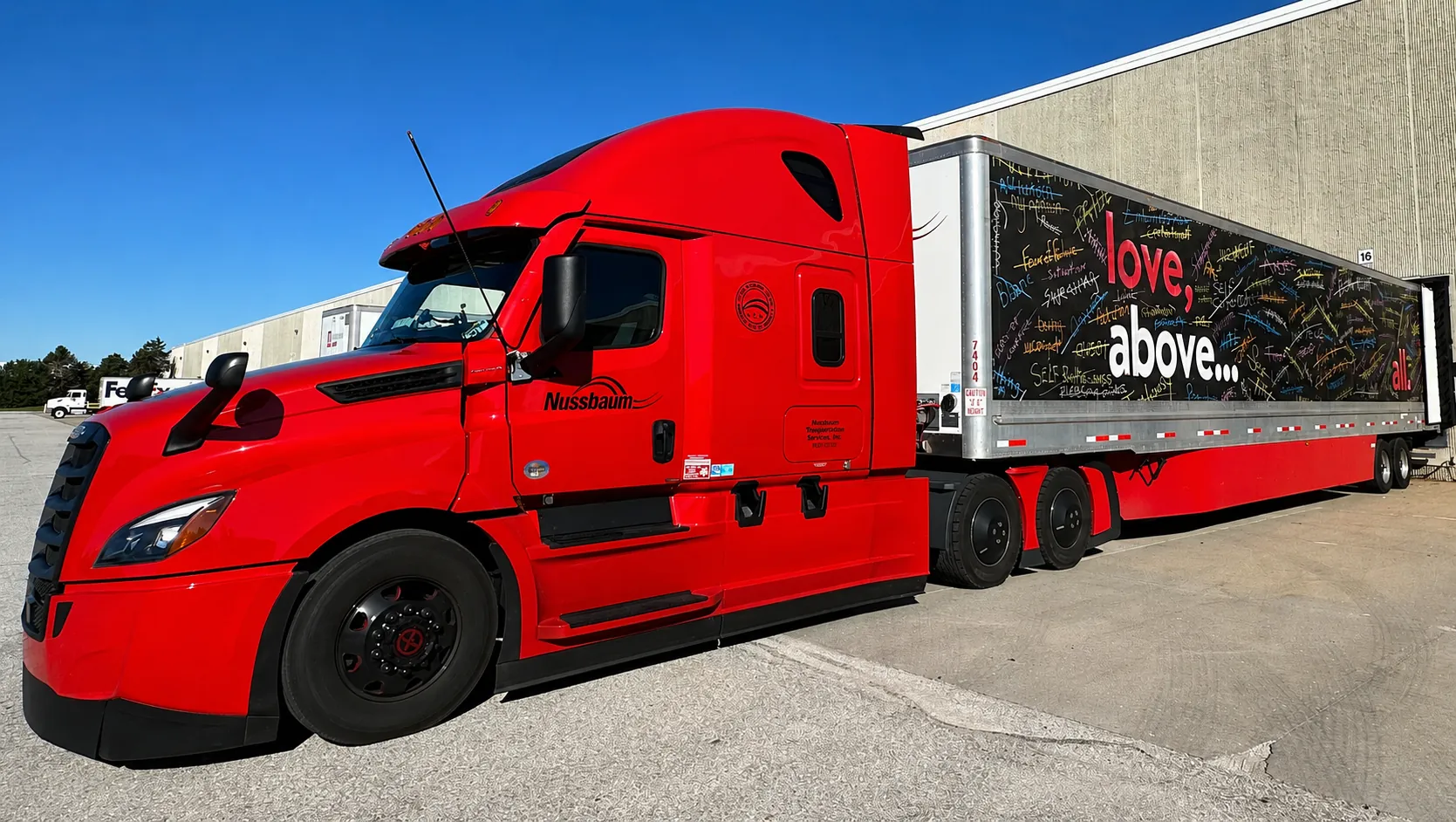Bright red tractor-trailer parked beside a warehouse under a blue sky